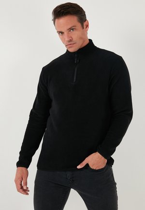 Schwarzer Fleece-Pullover mit hohem Kragen, viertelreißverschluss, langen Ärmeln und weicher Textur, getragen über dunklen Jeans.