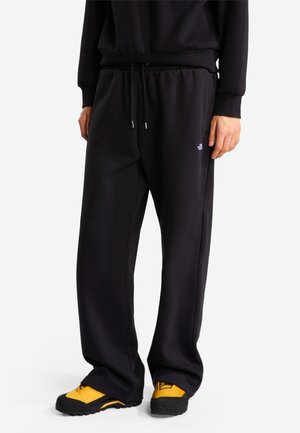 Man draagt zwarte losse joggingbroek met trekkoord en geel-zwarte buitenschoenen, staand tegen een witte achtergrond.