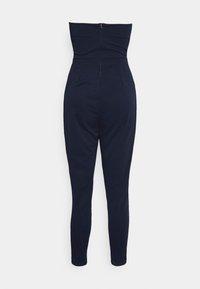 Macaquinho ajustado azul marinho com design de pernas estreitas, apresentando um topo estruturado, sem alças, e um fecho oculto nas costas. Textura suave.