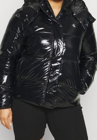 Czarna puchowa kurtka z błyszczącego materiału, z zapięciem na zamek błyskawiczny i zatrzask, posiadająca kieszenie naszywane oraz kaptur. Logo Calvin Klein na klatce piersiowej.