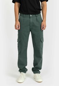 Green cargo pants made of cotton, featuring side pockets, a relaxed fit, and a smooth texture. Worn with white sneakers and a black t-shirt.