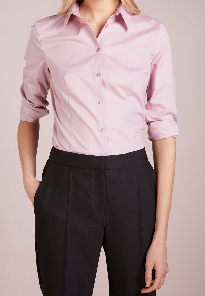 Personne portant une chemise rose pâle boutonnée avec les manches retroussées et un pantalon noir taille haute, les mains dans les poches.