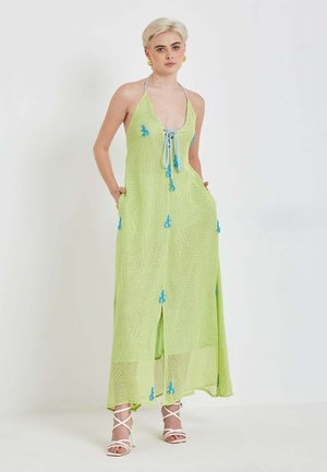 Mladi oseba s kratkimi svetlo blond lasmi, oblečena v svetlo zeleno maxi obleko brez rokavov z modrimi poudarki in belimi sandalami z visokimi petami, stoji.