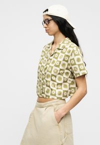 Γυναίκα με μπεζ καπέλο φοράει το καπέλο ανάποδα, γυαλιά με σκελετό χελώνας, πράσινο και μπεζ πουκάμισο με σύντομα μανίκια και φούστα μπεζ, με τα χέρια στις τσέπες.