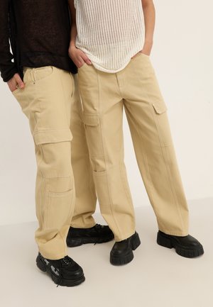 Beige cargo bukser med store lommer, synlige syningsdetaljer og en afslappet pasform, parret med klodsede sorte sko.