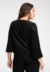 Czarna cekinowa bluzka z rękawami do połowy przedramienia, o luźnym kroju i fakturowanej powierzchni. Widok z tyłu podkreśla błyszczący materiał i drapowany fason.