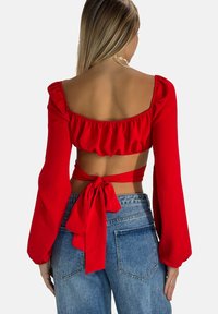 Crop top rosso con scollatura arricciata e maniche lunghe; legato sul retro. Abbinato a jeans di denim blu a vita alta. Texture liscia.