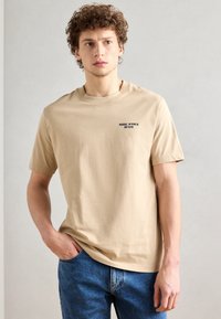 Tan bomulds t-shirt med kortærmer, der har et lille sort "Marc O'Polo Denim" logo på venstre bryst, parret med blå jeans.