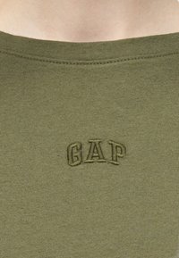 Ύφασμα χακί-πράσινο με κοντινή λήψη του κεντημένου λογότυπου "GAP" στο κέντρο κάτω από στρογγυλεμένη λαιμόκοψη.