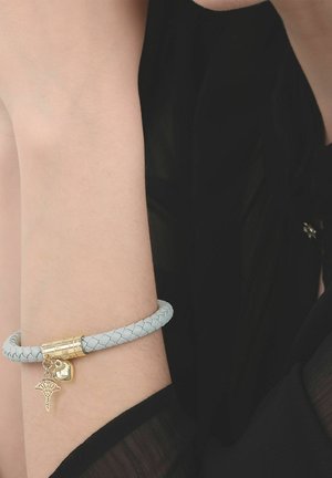 Muñeca con pulsera trenzada blanca que presenta dijes dorados en forma de corazón y árbol sobre un fondo de tela negra.