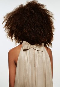 Ärmellös klänning i ljusguld med glansig struktur och rosettdetalj baktill. Har flödande tyg och hög halsringning.