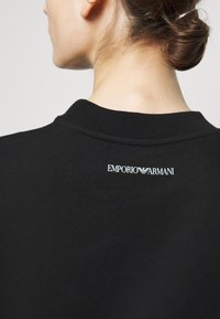 Felpa nera con collo a costine, con un piccolo logo bianco "EMPORIO ARMANI" centrato sulla schiena. Tessuto liscio.