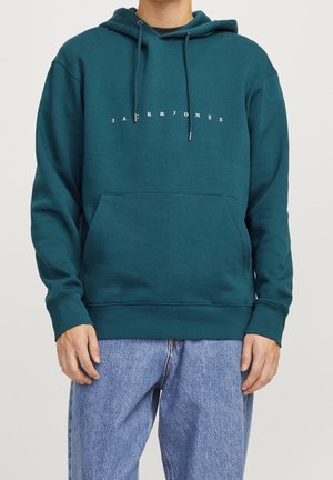 Osoba ubrana w turkusową bluzę z kapturem Jack & Jones z kieszenią z przodu i sznurkami, zestawioną z luźnymi jasnoniebieskimi jeansami.