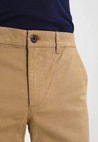 Pantalon en coton beige avec devant plat, fermeture à un bouton et poches latérales. Texture lisse avec des détails de couture visibles.