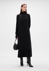 Μακρύ μαύρο πλεκτό φόρεμα με ψηλό γιακά τύπου turtleneck, ριμπ μανσέτες και χαλαρή σιλουέτα. Το μοντέλο κρατά μια μικρή μαύρη τσάντα και φοράει μπότες μέχρι το γόνατο.