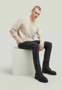 Maglione beige lavorato a maglia con motivi testurizzati, abbinato a jeans neri strappati e stivali neri in pelle stringati, seduto su un piedistallo bianco.