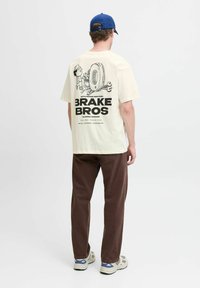 Cremefarvet t-shirt med et sort grafisk print, der forestiller en mekaniker, et dæk og værktøj på ryggen; parret med mørkebrune bukser.