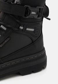 Gros plan d’une botte d’hiver noire isolante avec une semelle épaisse, une bande réfléchissante, une boucle sécurisée portant la mention « DM’s », et des détails de coutures renforcées.