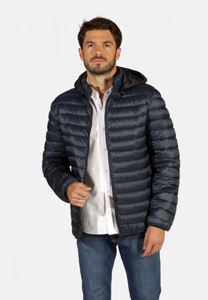 Doudoune marine avec un design matelassé, fermeture éclair et capuche intégrée. Tissu lisse et brillant ; portée sur une chemise blanche et un jean bleu.