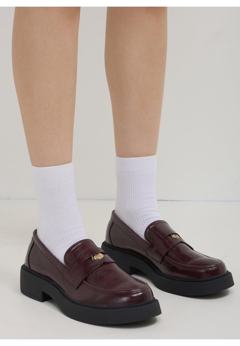 Jambes portant des chaussettes blanches côtelées et des mocassins en cuir bordeaux foncé avec des semelles épaisses noires et des détails dorés sur un fond uni.