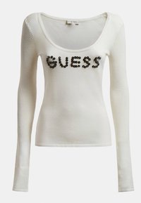 Maglietta bianca a maniche lunghe con scollo a barca, maniche in rete e borchie decorative nere che formano la parola "GUESS" sul davanti.