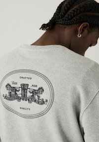 Sweat à capuche gris avec un grand logo circulaire au dos, orné d'illustrations noires de chevaux et de texte descriptif, en tissu doux et texturé.