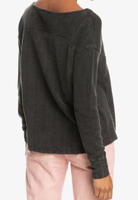 Persoon in een donkergrijs, getextureerd, langermanig shirt met verticale strepen en lichtroze broeken, gezien vanaf de achterkant.