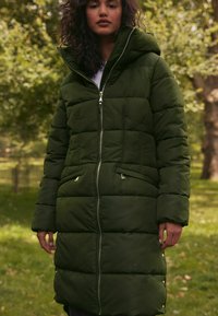 Zeleni puffa kaput s zatvaračem, velikim kapuljačom i quiltanim dezenom. Ima bočne džepove i metalne detalje.