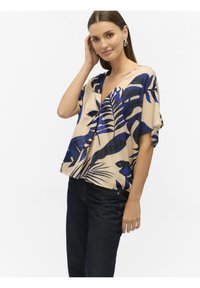 Crème kleurige blouse met blauwe tropische bladerenpatronen, relaxte pasvorm, korte mouwen, V-hals. Gecombineerd met donkerblauwe denim jeans.
