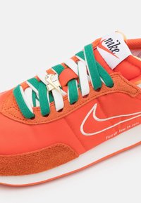 Orange sneaker med gröna och vita snören, mockadetaljer och en vit Nike-swoosh. Har en gulddetalj och präglad text.
