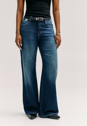 Persoon die wijdvallende donkerblauwe jeans draagt met een zwarte riem, zwart mouwloos topje, zwarte schoenen en een zilveren armband aan de rechterpols.