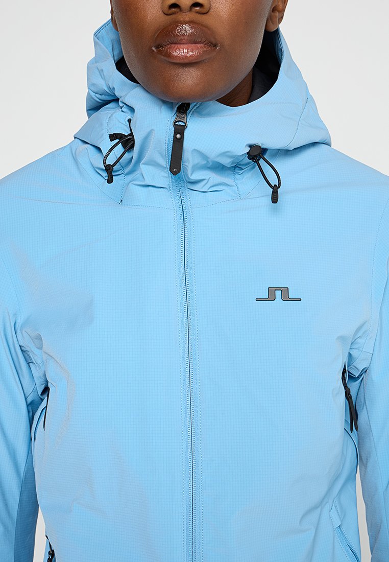 Jasnoniebieska wodoodporna kurtka o dopasowanym kroju, z kapturem i zamkami. Posiada delikatny wzór w kratkę i czarne akcenty sprzętowe.