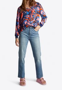 Kolorowa bluzka z długim rękawem z wirującymi różowymi, niebieskimi i czerwonymi wzorami, w połączeniu z jasnoniebieskimi prostymi dżinsami i różowymi sandałami.