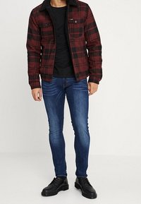 Červená a černá károvaná bunda s černým umělým kožešinovým límcem, doplněná černou košilí, tmavě modrými úzkými džínami a černými společenskými botami.