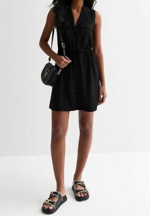 Žena v černých bezrukávových šatech na knoflíky s mašlí v pase, nesoucí černou kabelku přes rameno a černé sandály s řetízkovými detaily.