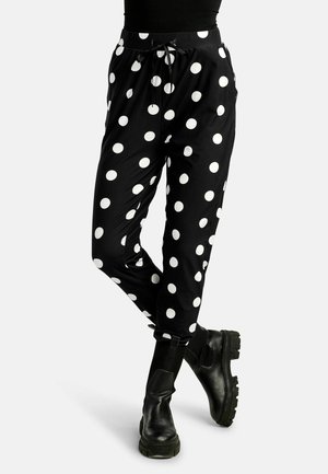 Nogi w czarnych spodniach w duże białe grochy, zestawione z masywnymi czarnymi botkami na jasnym tle.