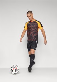 Sort fodboldtrøje med orange accenter, der har et bølgende rødt mønster. Kombineret med sorte shorts, sorte strømper og fodboldsko, ved siden af en bold.