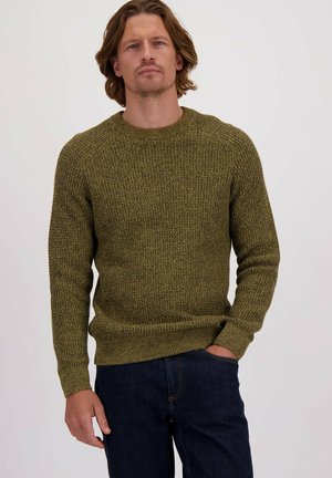 Man met gemiddeld lang haar, gekleed in een olijfgroene gebreide trui met textuur en donkerblauwe jeans, met één hand in zijn zak.