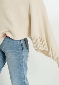 Poncho de punto beige con flecos largos en el dobladillo, llevado sobre jeans azules. La tela parece suave con un acabado sutil y texturizado.