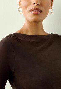 Ruskea pitkähihainen paita, jossa on veneen muotoinen pääntie. Olkapäissä on rypytyst yksityiskohtia. Mallilla on päällä kultaketjuiset korvakorut. Tasainen pinta.