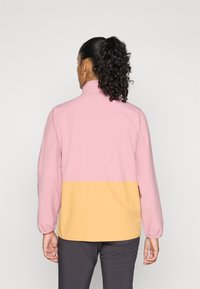 Mujer vista de espaldas llevando una chaqueta de bloques de color rosa y amarillo, con mangas largas y cuello alto, de pie frente a un fondo sencillo.