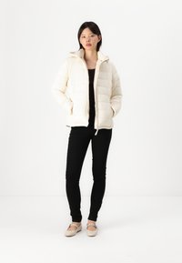 Aufblasbare Jacke in Off-White, mit einem gesteppten Design, Kapuze und Reißverschluss. Kombiniert mit schwarzen Hosen und beigen Schuhen.