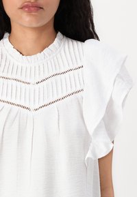 Blouse blanche avec un tissu texturé, des manches à volants, des détails froncés et des découpes sur le haut devant.