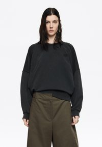 Čierna oversize mikina s raglánovými rukávmi, vyrobená z mäkkého materiálu, s jemným logom na hrudi. V kombinácii s olivovozelenými nohavicami.
