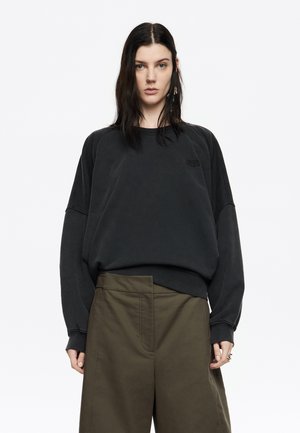 Črna oversize trenerka z raglan rokavi, narejena iz mehkega materiala, z diskretnim logotipom na prsih. V kombinaciji z olivno zelenimi hlačami.