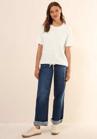 T-shirt blanc avec une poche poitrine et un ourlet à cordon, associé à un jean bleu ample retroussé et des baskets beiges. Texture lisse et design décontracté.