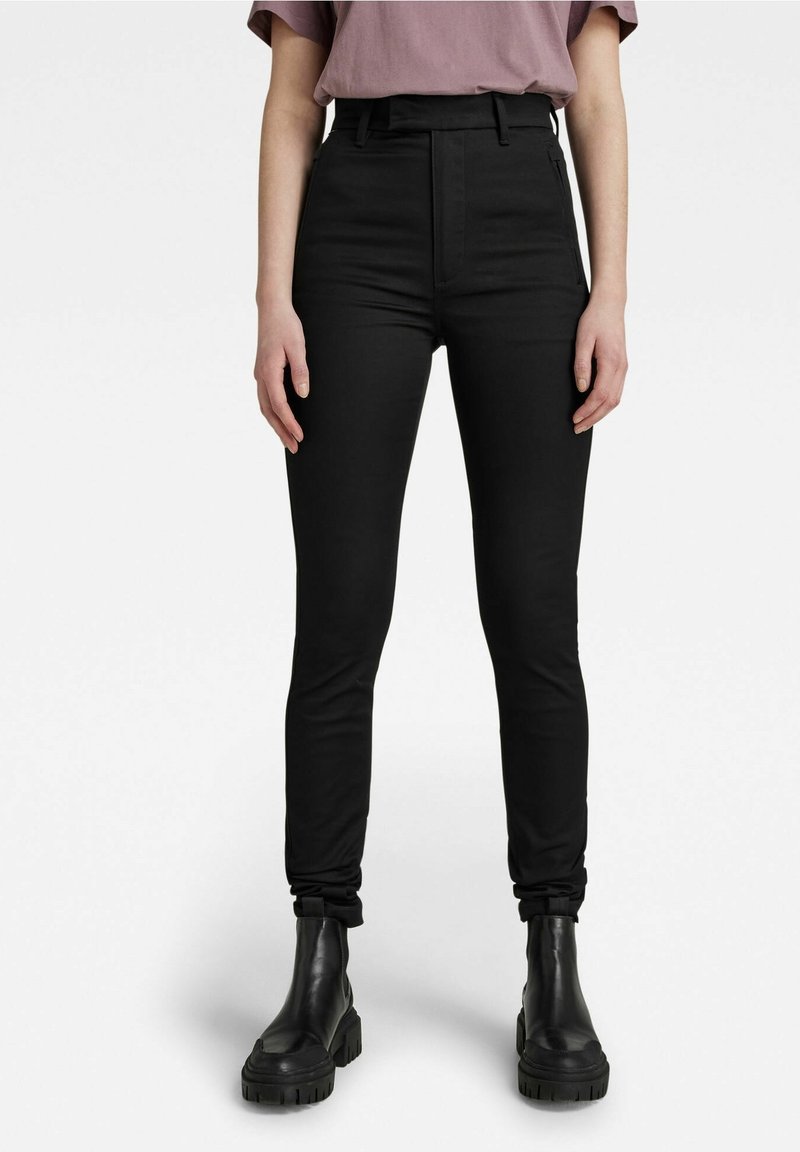 Jean skinny noir taille haute en tissu extensible, avec une fermeture éclair à l'avant et des poches latérales discrètes, assorti à des bottines noires.