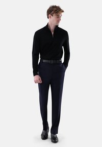 Schwarzer Reißverschluss-Pullover, maßgeschneiderte marineblaue Hose und schwarze Schuhe. Das Ensemble zeichnet sich durch eine schlanke Passform mit glatten Texturen und minimalen Details aus.