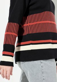Schwarzer Pullover mit auffälligen roten horizontalen Streifen, schmalen vertikalen Linien und weißen Akzenten an den Bündchen, aus einem weichen Strickmaterial.