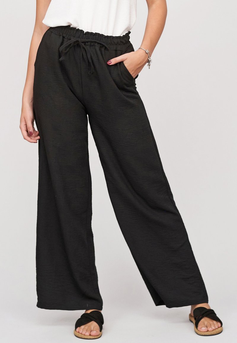 Femme portant un pantalon noir à jambes larges avec taille à cordon, un haut blanc et des sandales noires à brides croisées, debout avec une main dans la poche.